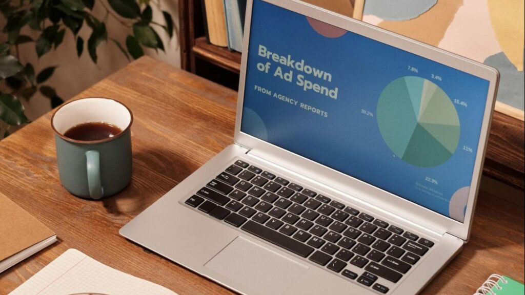 A laptop on top of a brown table with coffee at the side displaying "Breakdown of Ad Spend" in their paid ads.