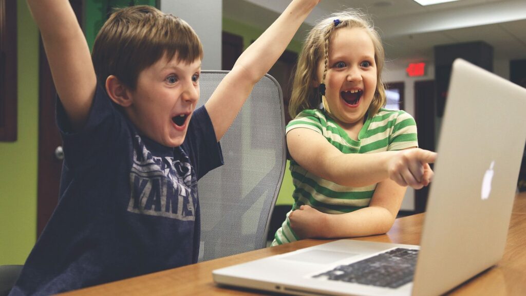 Two kids celebrating in front of their laptop while the girl is pointing on the laptop screen.