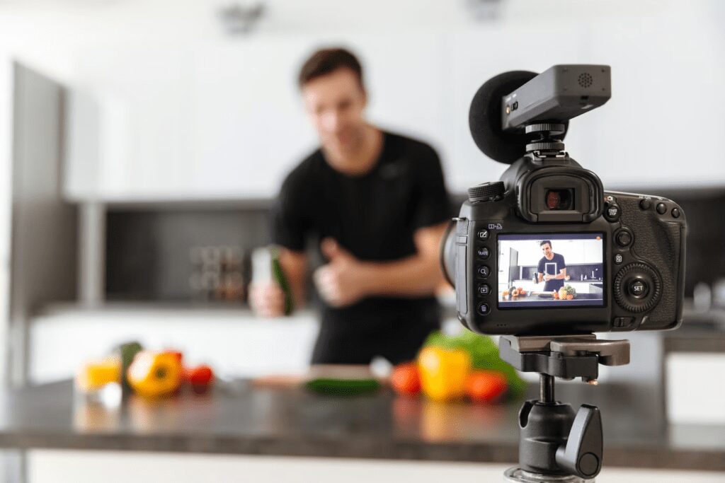 A man in black shirt in their kitchen with various fruits while they are in front of the video camera.