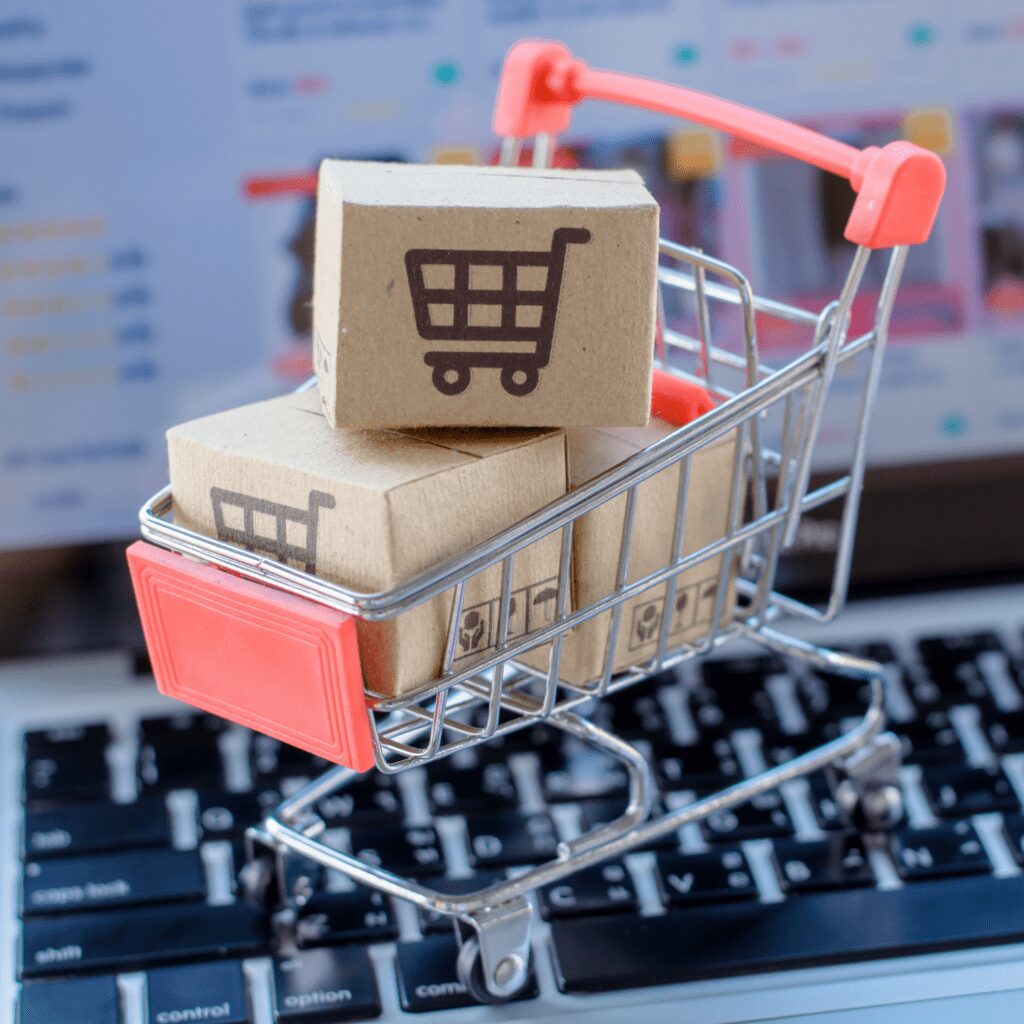 A mini grocery cart filled with small carton boxes labeled with grocery carts too while being on top of a laptop to represent marketing.